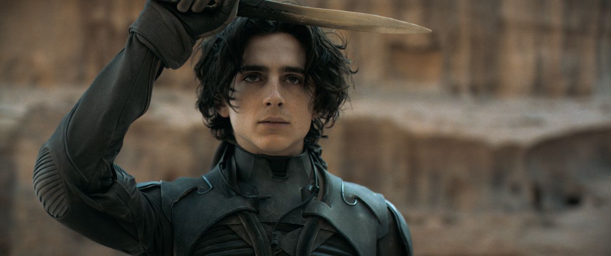 Timothée Chalamet as Paul Atreides holding up a dagger in a desert valley in Dune