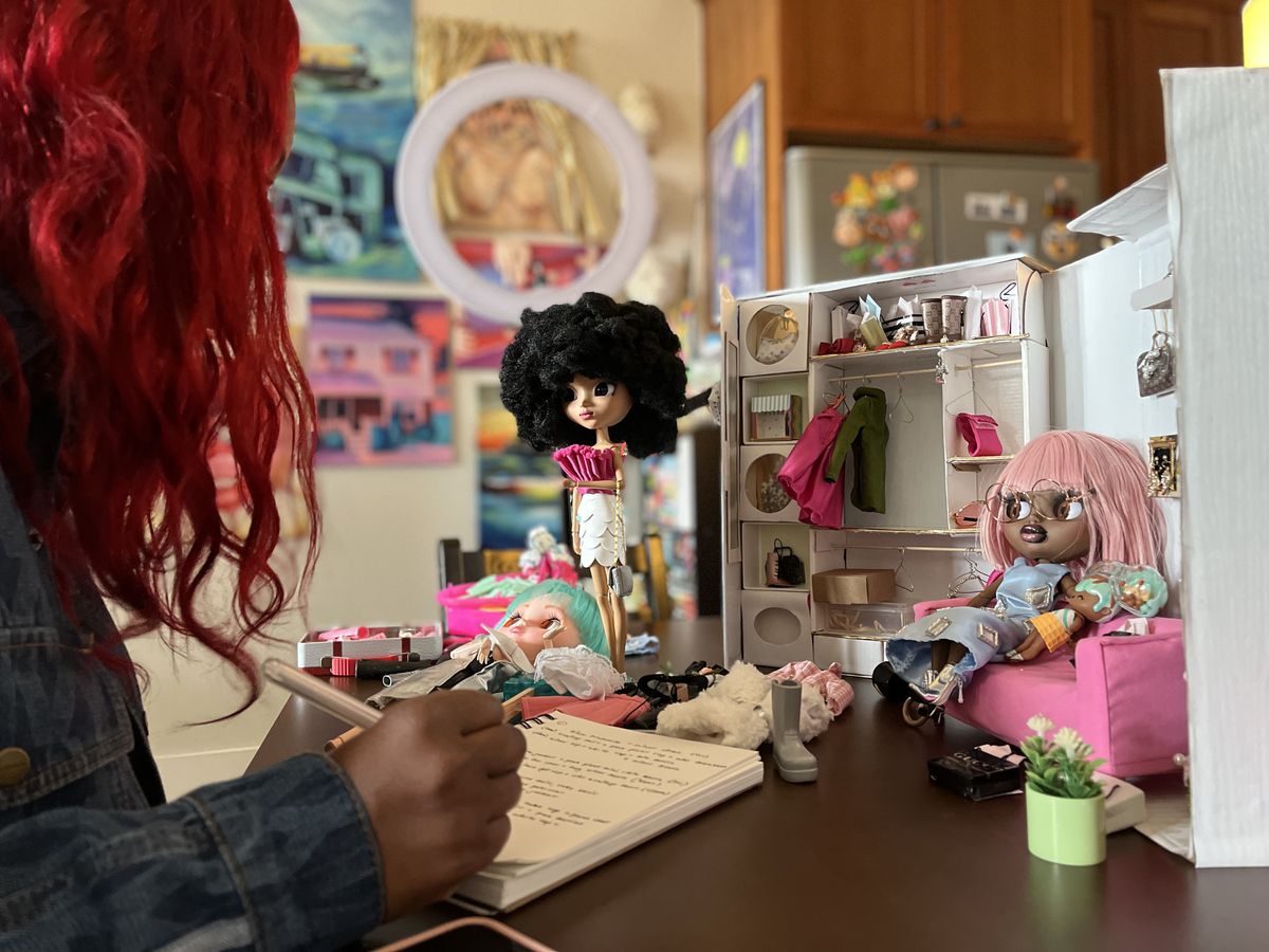 A redhaired woman writes on a pad of paper in front of two Blythe dolls set up in a small bedroom diorama