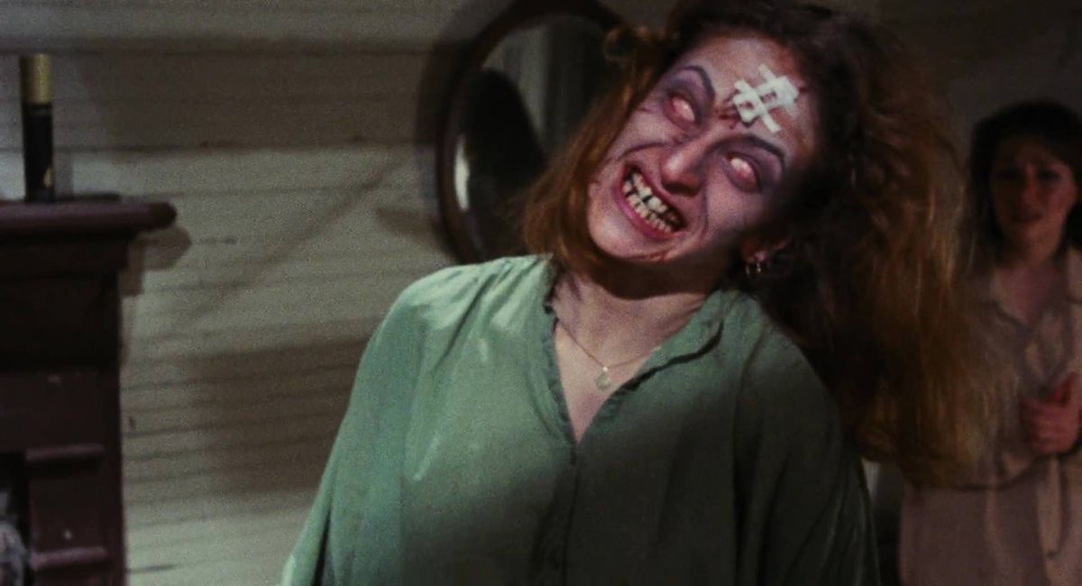 A young woman starts to transform into a Deadite, eyes white, with a mischievous smile on her face, in The Evil Dead.